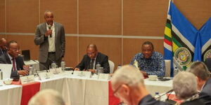 Former Foreign PS Macharia Kamu (standing) addressing a delegation in presence of former President Uhuru Kenyatta (right).