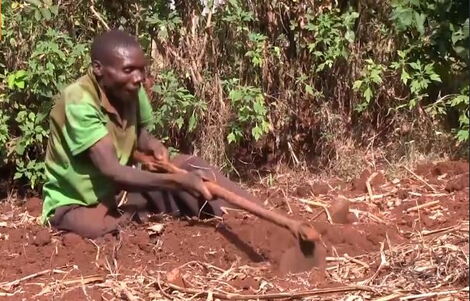 An Image of Simon Chege Digging a Piece of Land While Seated